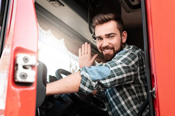 Young smiling male truck driver sitting inside truck and waving with his hand