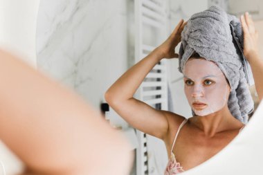 Banyoda yüzünde uygulamalı çarşaf maskesi olan güzel bir kadın aynaya bakıyor.