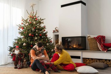 Genç ve mutlu bir aile, Noel ağacı ve şenlikli süslemelerle süslenmiş sıcak bir oturma odasında, parlayan bir şöminenin yanında oturuyor..