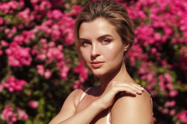Yumuşak tenli ve doğal makyajlı güzel sarışın kadın narin pembe çiçeklerle dolu bir çalılığın arkasında poz veriyor..