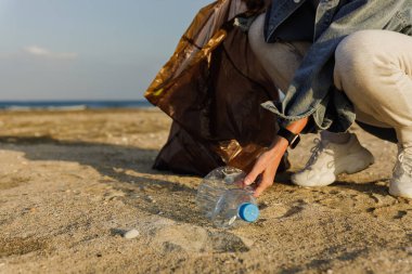 Kadın gönüllü, doğayı temiz tutma çabalarına katkıda bulunmak için plajda plastik atık topluyor. Plastik şişeyle yakın plan..
