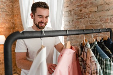 Genç adam gardıroptaki elbise rafında asılı çeşitli gömleklerini karıştırıyor.