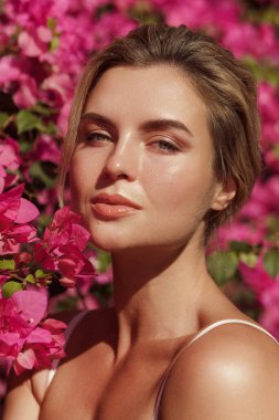 Yumuşak tenli ve doğal makyajlı güzel sarışın kadın narin pembe çiçeklerle dolu bir çalılığın arkasında poz veriyor..