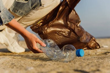 Kadın gönüllü, doğayı temiz tutma çabalarına katkıda bulunmak için plajda plastik atık topluyor. Plastik şişeyle yakın plan..