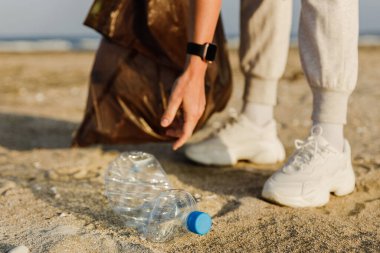 Kadın gönüllü, doğayı temiz tutma çabalarına katkıda bulunmak için plajda plastik atık topluyor. Plastik şişeyle yakın plan..