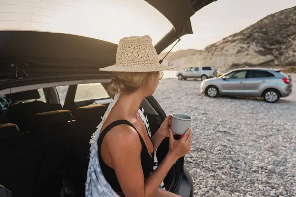 Traveler woman taking a break with coffee by her car on a pebble beach.