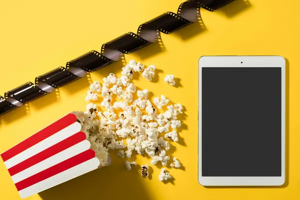 Classic popcorn bucket and tablet computer with blank screen on yellow background.