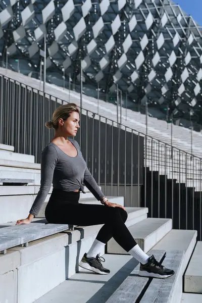 Woman athlete wearing female sportswear sitting on bench of bleachers in outdoor stadium.