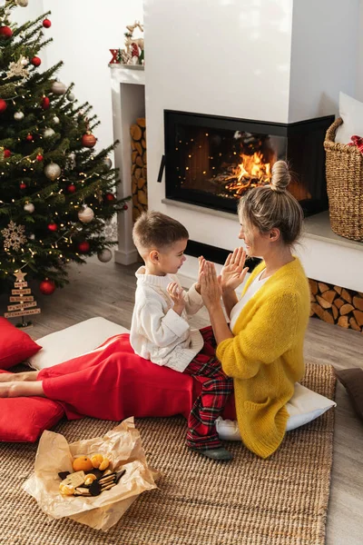 女性と彼女の小さな息子は クリスマスツリーとお祝いの装飾で飾られた居心地の良いリビングルームで輝く暖炉の横に手拍子ゲームをプレイします — ストック写真