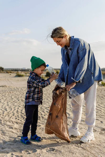 コーカサス人の家族はビーチでプラスチック廃棄物を集めています 母は自然をきれいに保つのを助けるために息子に教えています — ストック写真