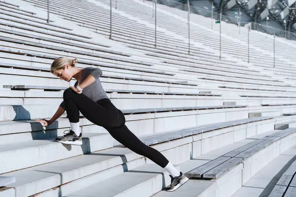 Spor giyinen kadın sporcu açık hava stadyumunun merdivenlerinde bacak esnetiyor..