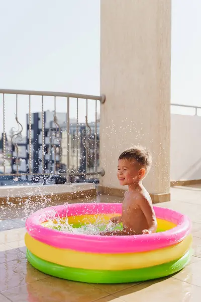 Toddler boy gleefully plays in an inflatable round swimming pool on the balcony during a hot summer day.