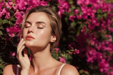 Yumuşak tenli ve doğal makyajlı güzel sarışın kadın narin pembe çiçeklerle dolu bir çalılığın arkasında poz veriyor..