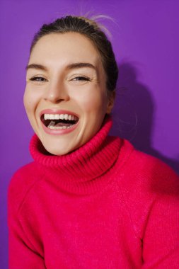 Geniş açılı, mor arka planda sıcak polo yaka kazak giyen neşeli bir kadının yakın plan portresi.