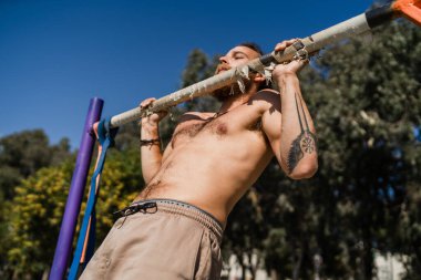 Genç ve kaslı bir adam sokak antrenmanı sırasında barfiks çekiyor.