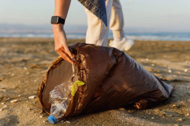Kadın gönüllü, doğayı temiz tutma çabalarına katkıda bulunmak için plajda plastik atık topluyor. Plastik şişeyle yakın plan..