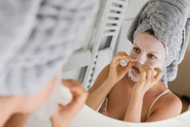 Banyoda yüzünde uygulamalı çarşaf maskesi olan güzel bir kadın aynaya bakıyor.