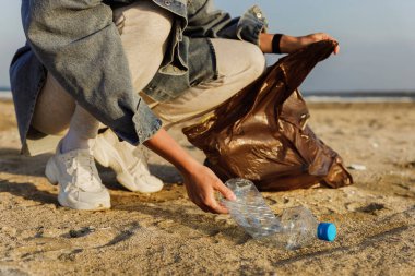 Kadın gönüllü, doğayı temiz tutma çabalarına katkıda bulunmak için plajda plastik atık topluyor. Plastik şişeyle yakın plan..