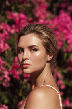 Yumuşak tenli ve doğal makyajlı güzel sarışın kadın narin pembe çiçeklerle dolu bir çalılığın arkasında poz veriyor..