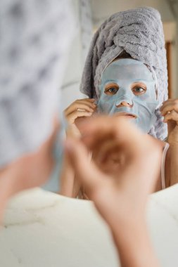 Banyoda yüzünde uygulamalı çarşaf maskesi olan güzel bir kadın aynaya bakıyor.