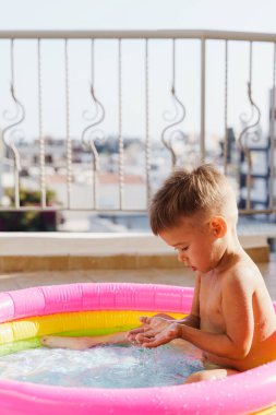 Küçük çocuk sıcak bir yaz günü balkonda şişirilebilir yuvarlak bir yüzme havuzunda neşeyle oynar..