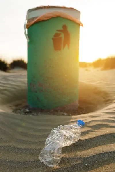 Plastic bottle near green waste container on sandy beach. Plastic waste pollution and greenwashing.