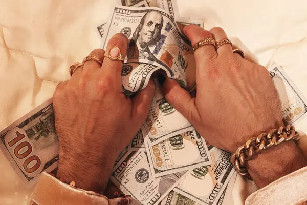 Hands of a wealthy man, adorned with golden rings and a striking black manicure against a backdrop of US dollar bills.