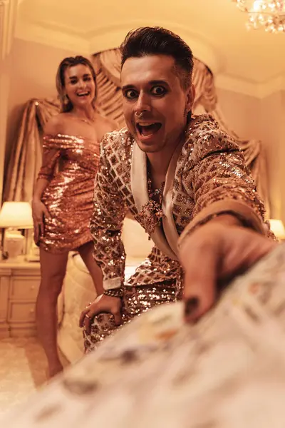 Wealthy young couple is dressed in radiant golden outfits adorned with sequin embroidery, situated in a luxurious suite.