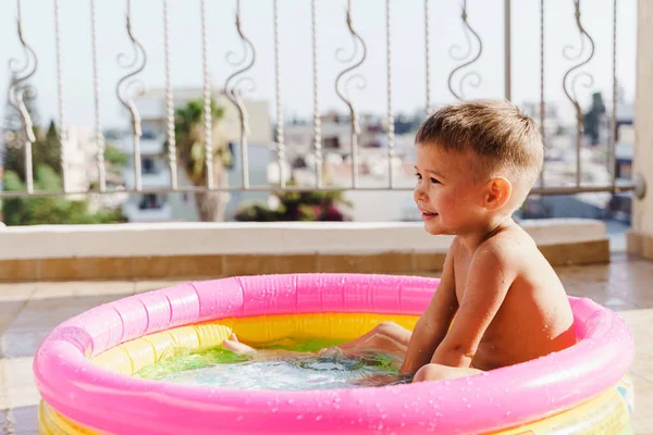 Toddler boy gleefully plays in an inflatable round swimming pool on the balcony during a hot summer day.