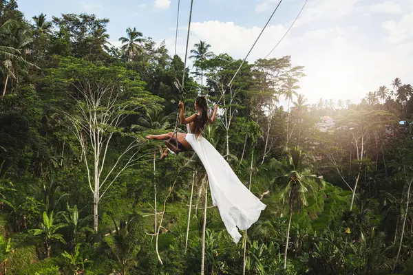 stock image Young woman wearing white dress swinging on rope swings with beautiful view on rice terraces and palm trees in the Bali Island