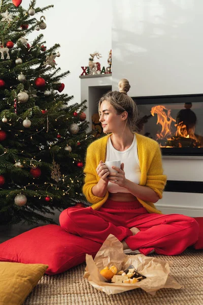 幸せな女性は居心地の良いリビングルームに輝く暖炉の横に座り クリスマスツリーとお祝いの装飾で飾られ ホットドリンクを楽しんでいます — ストック写真