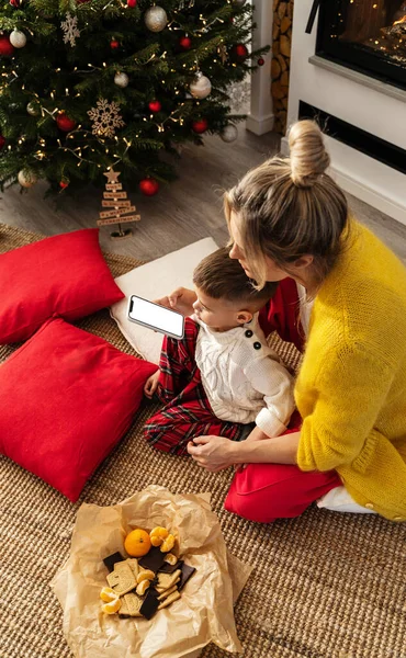 女性と彼女の小さな息子は クリスマスツリーとお祝いの装飾で飾られた居心地の良いリビングルームで空白のスクリーンでスマートフォンを見ます — ストック写真