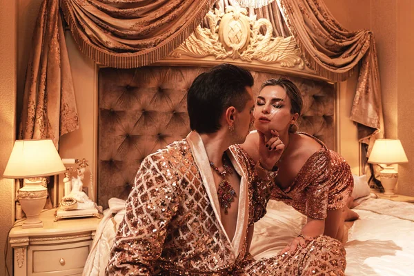 Wealthy young couple is dressed in radiant golden outfits adorned with sequin embroidery, situated in a luxurious suite.