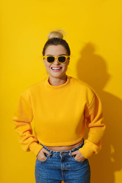 Portrait of young cheerful girl wearing yellow sweatshirt and sunglasses against yellow background