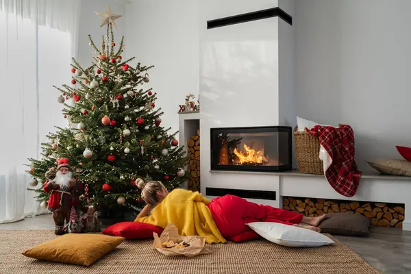 若い女性は居心地の良いリビングルームの輝く暖炉の横にあり クリスマスツリーとお祝いの装飾で飾られています — ストック写真