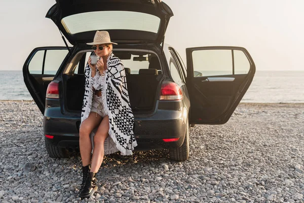 Traveler woman taking a break with coffee by her car on a pebble beach.