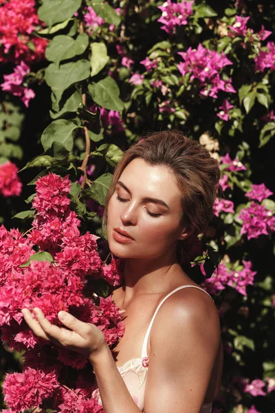 Beautiful blonde woman with smooth skin and natural makeup, as she poses against a backdrop of a bush with delicate pink flowers.