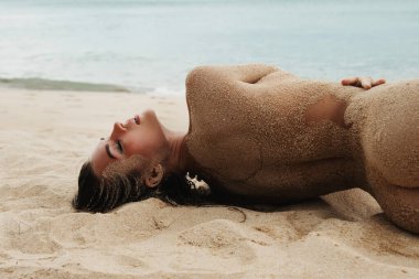 Muhteşem genç bir kadın kumsalda, vücudu ustalıkla kum taneleriyle kaplıydı..
