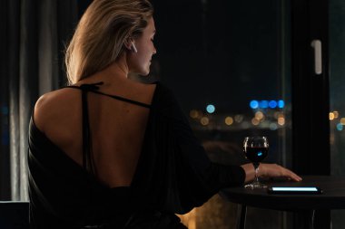 Gece vardiyasında yalnız bir kadın, akıllı telefonuyla meşgul kırmızı şarap yudumluyor ve kablosuz kulaklık takıyor..