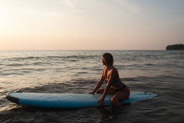 Kadın gün batımında okyanusta sörf tahtasında oturuyor..