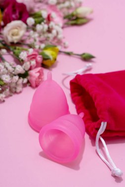 Pembe arka planda canlı, taze çiçeklerin bulunduğu bir koleksiyonun yanında, çevre dostu bir dişi ürünü tasvir eden regl bardakları..