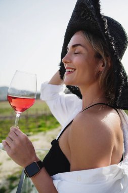 Zarif giyinmiş ve geniş kenarlı şapkalı bir kadın üzüm bağında bir bardak gül şarabı içiyor.