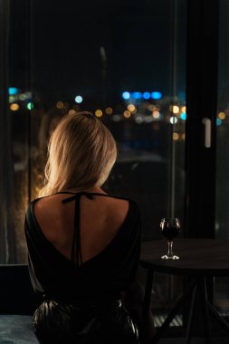 Gece vakti iç mekanda yalnız bir kadın, sakince pencereden dışarı bakarken bir bardak kırmızı şarabın tadını çıkarıyor..