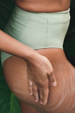 Bikinili bir kadının, gür tropikal yapraklarla çevrili kalçasına losyon sürerken yakın çekimi..