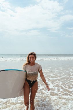 Neşeli kadın sahilde yürürken mavi bir sörf tahtası taşıyor.. 