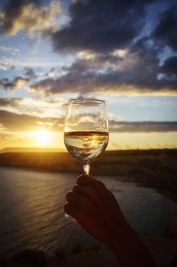 Bir bardak beyaz şarapla kadın eli, nefes kesici bir deniz batımına karşı..