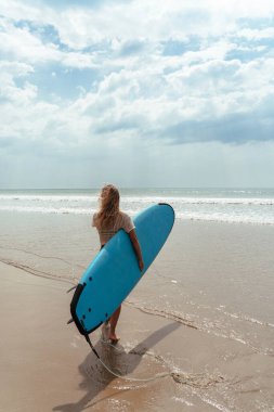 Neşeli kadın sahilde yürürken mavi bir sörf tahtası taşıyor..