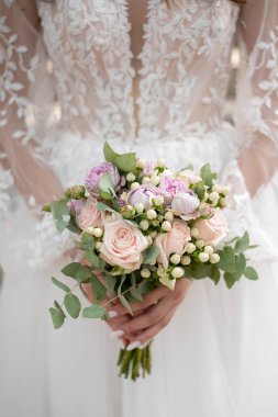 Düğün buketi gelinin elinde. Güller beyaz bir elbisenin arka planında bir buket içinde toplanır. Düğün ajansları ve düğün hizmetleri konsepti. nakışlı kumaş ve kollu desenler