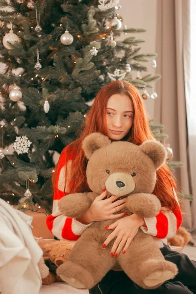 Happy redhead woman in sweater with toy bear under Christmas tree with lights. Christmas