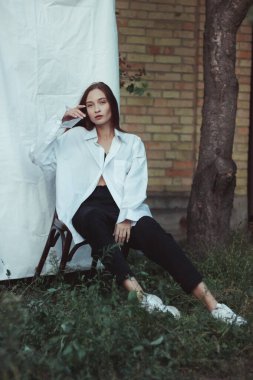 Beyaz gömlekli bir kızın modern fotoğrafı beyaz bir çarşafa karşı. Sandalyede poz vermek. Kendine güvenen bir kadının lakonik fotoğrafları. Bitki arkaplanı